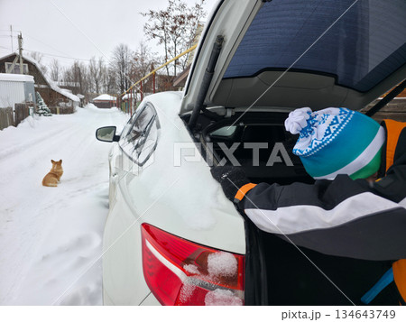 Man cleaning snow from car trunk in winter village street, with a dog sitting nearby. Cold season and travel preparation. 134643749