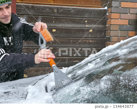 Man removing snow from car windshield with an ice scraper and brush. Winter car maintenance and preparation for cold weather. Man removing snow from car windshield with an ice scraper and brush. Winter car maintenance and preparation for cold weather. 134643877