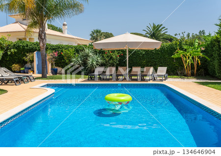 Families enjoy a sunny afternoon by the pool, featuring lounge chairs and a large umbrella in a vibrant garden filled with greenery 134646904