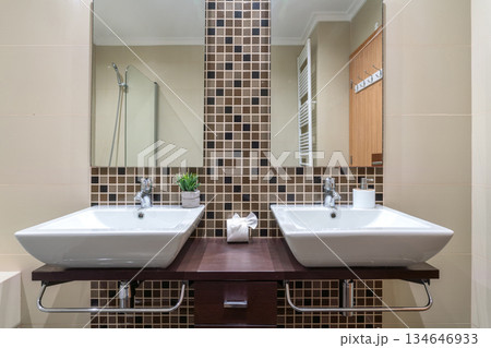 Stylish bathroom showcases two white sinks on a wooden counter, complemented by square mosaic tiles and double mirrors, creating a contemporary look Stylish bathroom showcases two white sinks on a wooden counter, complemented by square mosaic tiles and double mirrors, creating a contemporary look 134646933