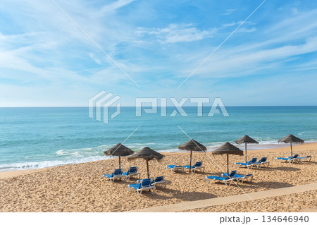 Sun-kissed sandy beach features lounge chairs arranged beneath thatched umbrellas, inviting relaxation by the calm ocean waves and clear blue skies Sun-kissed sandy beach features lounge chairs arranged beneath thatched umbrellas, inviting relaxation by the calm ocean waves and clear blue skies 134646940