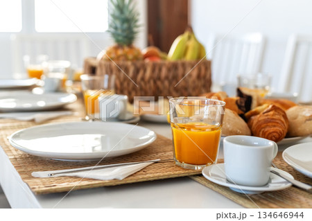 A table is set for breakfast featuring orange juice, fresh fruit, and a variety of pastries. Natural light fills the room, creating a warm, inviting atmosphere A table is set for breakfast featuring orange juice, fresh fruit, and a variety of pastries. Natural light fills the room, creating a warm, inviting atmosphere 134646944