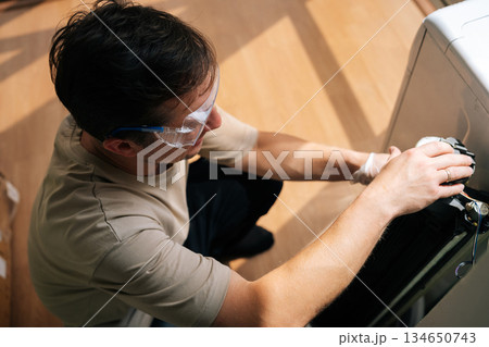 Top view of repairman wearing safety glasses using screwdriver diligently fixes broken washing machine, providing essential maintenance services for home appliances with expertise and care. Top view of repairman wearing safety glasses using screwdriver diligently fixes broken washing machine, providing essential maintenance services for home appliances with expertise and care. 134650743