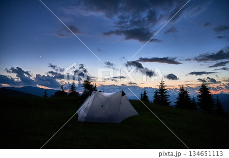 Touristic tent glows warmly on grassy hillside at dusk, surrounded by dark silhouettes of mountains and pine trees. Sky is deep blue with scattered clouds, as last light of sunset fades into night. 134651113
