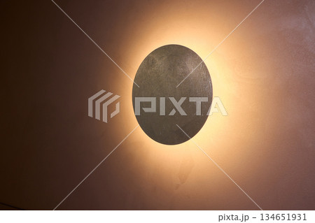 Elegant textured fixture casting gentle golden glow, Circular textured sconce mounted on plaster wall emitting soft gold light and subtle radial shadows for intimate setting 134651931