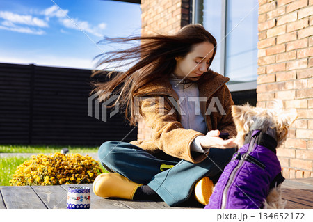 A happy young woman sitting cross-legged outdoors with her small dog on a sunny day, cozy autumn outfit and calm lifestyle scene, pet human friendship 134652172