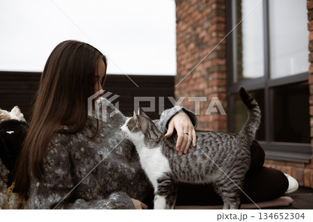 Warm autumn moment of human and animal connection. A young woman in cozy knitted sweater sitting on patio, playing with a cat, enjoying a calm day. 134652304
