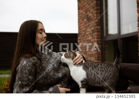 Warm autumn moment of human and animal connection. A young woman in cozy knitted sweater sitting on patio, playing with a cat, enjoying a calm day. 134652305