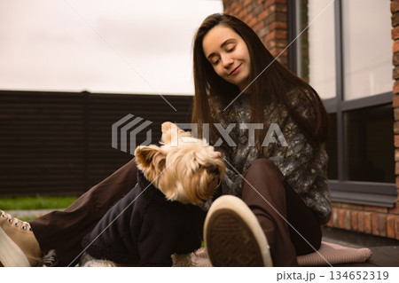 A young pretty white woman sitting on patio near a modern brick house, smiling at her small Yorkshire Terrier dog wearing a jacket. Human and pet. A young pretty white woman sitting on patio near a modern brick house, smiling at her small Yorkshire Terrier dog wearing a jacket. Human and pet. 134652319