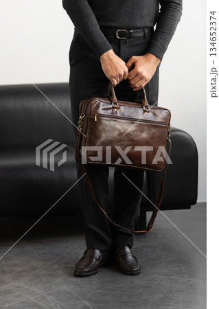 A professional man in a charcoal crewneck sweater and tailored trousers holds a brown leather briefcase standing in a minimalist studio. A professional man in a charcoal crewneck sweater and tailored trousers holds a brown leather briefcase standing in a minimalist studio. 134652374