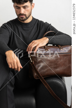 A professional male model in charcoal knitwear and tailored trousers sits on a black leather sofa holding a premium brown leather briefcase. A professional male model in charcoal knitwear and tailored trousers sits on a black leather sofa holding a premium brown leather briefcase. 134652379