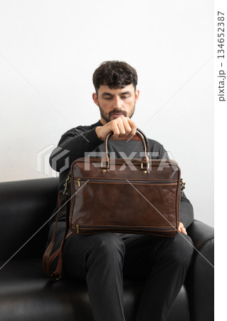 A professional male model in charcoal knitwear and tailored trousers sits on a black leather sofa holding a premium brown leather briefcase. A professional male model in charcoal knitwear and tailored trousers sits on a black leather sofa holding a premium brown leather briefcase. 134652387