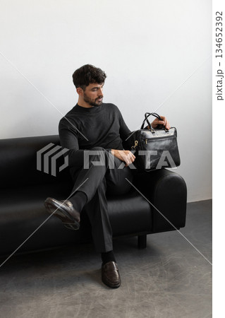 A professional male model in charcoal knitwear and tailored trousers sits on a black leather sofa holding a premium black leather briefcase. A professional male model in charcoal knitwear and tailored trousers sits on a black leather sofa holding a premium black leather briefcase. 134652392