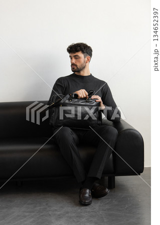 A professional male model in charcoal knitwear and tailored trousers sits on a black leather sofa holding a premium black leather briefcase. A professional male model in charcoal knitwear and tailored trousers sits on a black leather sofa holding a premium black leather briefcase. 134652397