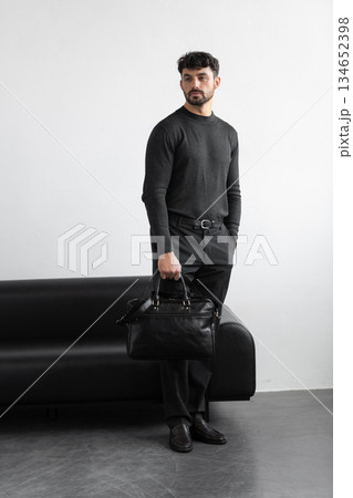 A professional man in a charcoal crewneck sweater and tailored trousers holds a black leather briefcase standing in a minimalist studio. A professional man in a charcoal crewneck sweater and tailored trousers holds a black leather briefcase standing in a minimalist studio. 134652398