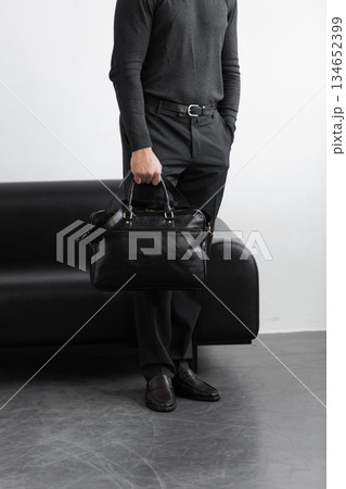 A professional man in a charcoal crewneck sweater and tailored trousers holds a black leather briefcase standing in a minimalist studio. 134652399