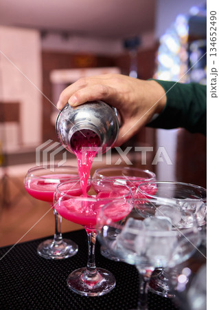 Mood setting with lively drink preparation at upscale bar, Elegant scene of bartender pouring colorful cocktail amidst festive warm lighting and guests in background Mood setting with lively drink preparation at upscale bar, Elegant scene of bartender pouring colorful cocktail amidst festive warm lighting and guests in background 134652490