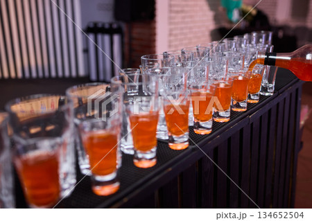 Celebratory drink scene, Orange liqueur shots at bar, Bartender pours drinks amidst lively party ambiance, Glassware reflects warm light during festive celebration at bar 134652504