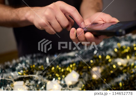 Digital holiday planning, Person reviewing online presents on tabletop, Warm indoor scene of individual managing digital gift list amidst festive lights and ornaments Digital holiday planning, Person reviewing online presents on tabletop, Warm indoor scene of individual managing digital gift list amidst festive lights and ornaments 134652778