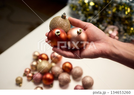 Hands arranging Christmas New Year baubles on white table with tinsel and warm bokeh lights, metallic red gold pink palette, careful styling by home decorator, festive craft mood Hands arranging Christmas New Year baubles on white table with tinsel and warm bokeh lights, metallic red gold pink palette, careful styling by home decorator, festive craft mood 134653260