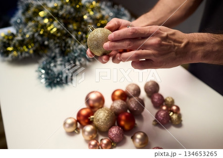 Hands arranging Christmas New Year baubles on white table with tinsel and warm bokeh lights, metallic red gold pink palette, careful styling by home decorator, festive craft mood 134653265