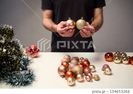 Hands arranging Christmas New Year baubles on white table with tinsel and warm bokeh lights, metallic red gold pink palette, careful styling by home decorator, festive craft mood 134653268