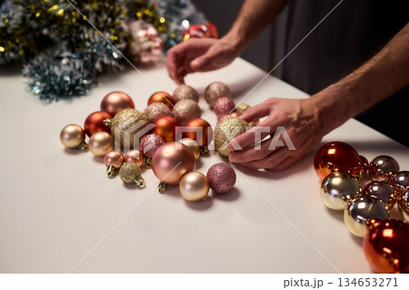 Hands arranging Christmas New Year baubles on white table with tinsel and warm bokeh lights, metallic red gold pink palette, careful styling by home decorator, festive craft mood 134653271