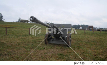 field howitzer positioned on open grass with long barrel and carriage visible, countryside houses on horizon give context to decommissioned training ground turned memorial display under muted sky. 134657765
