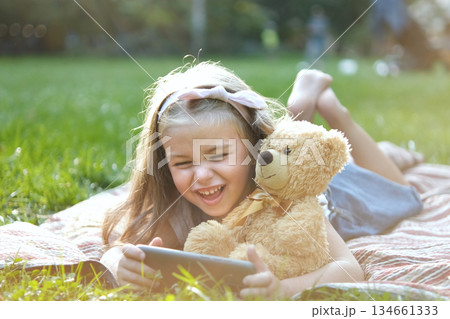 Small kid girl looking in her mobile phone together with her favorite teddy bear toy outdoors in summer park. Small kid girl looking in her mobile phone together with her favorite teddy bear toy outdoors in summer park. 134661333