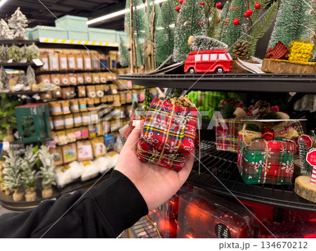 Person's hand holding a festive red plaid Christmas gift box, selecting charming holiday decorations for seasonal celebration in a bustling retail store 134670212