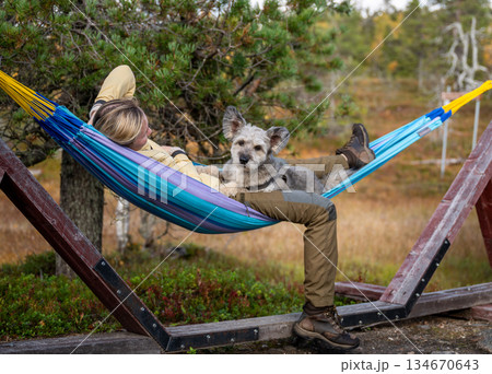 Woman relaxing in hammock with dog wearing outdoor clothing in forest nature Woman relaxing in hammock with dog wearing outdoor clothing in forest nature 134670643