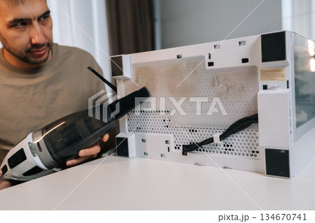 Focused man cleaning computer system unit with vacuum cleaner, removing dust from dirty air filter and mesh panel, providing essential pc maintenance and hardware care for optimal performance. 134670741