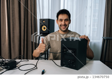 Portrait of young man smiling looking camera and giving thumbs up gesture, connecting studio monitor speaker for home recording or music production setup, expressing satisfaction with audio equipment. Portrait of young man smiling looking camera and giving thumbs up gesture, connecting studio monitor speaker for home recording or music production setup, expressing satisfaction with audio equipment. 134670750
