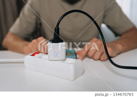 Cropped shot of person wirelessly setting up smart socket device plugged into power strip, interacting with smartphone display for home automation and remote control of appliances. 134670765