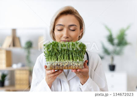 A female doctor in a white coat holds and smells a tray of fresh microgreens, possibly for nutritional assessment. 134672023