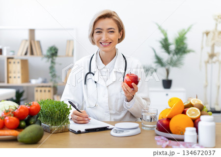 A smiling nutritionist holds an apple, surrounded by fresh fruits and vegetables, promoting healthy eating habits. A smiling nutritionist holds an apple, surrounded by fresh fruits and vegetables, promoting healthy eating habits. 134672037