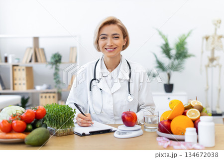 A smiling nutritionist sits at a desk surrounded by fresh fruits and vegetables, ready to consult on healthy eating. 134672038