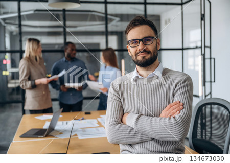 Man is standing with arms crossed. Professional workers are in the modern office together Man is standing with arms crossed. Professional workers are in the modern office together 134673030