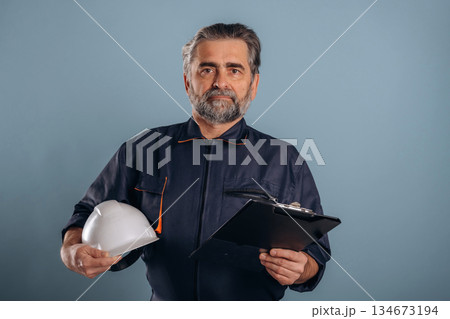 White hardhat and notepad. Senior man is standing in the studio against background White hardhat and notepad. Senior man is standing in the studio against background 134673194