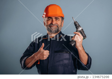 Repairer, holding drill, in hardhat. Senior man is standing in the studio against background 134673200