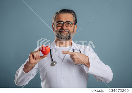 Stethoscope on neck, holding little heart. Senior man is standing in the studio against background 134673260