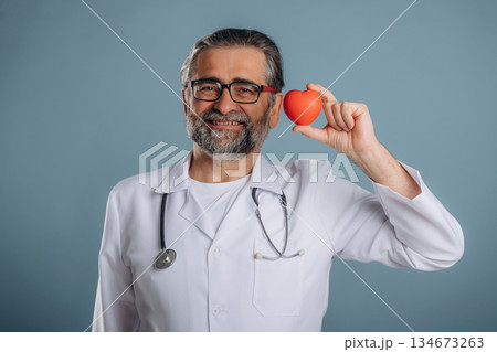 Stethoscope on neck, holding little heart. Senior man is standing in the studio against background 134673263