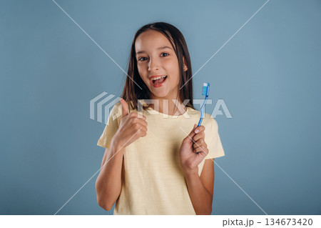 Thumb up, smiling and holding toothbrush. Young girl is against background Thumb up, smiling and holding toothbrush. Young girl is against background 134673420