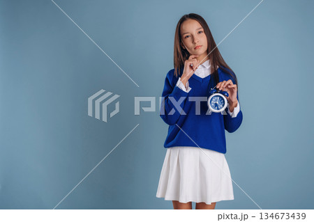 With blue clock in hand. Young schoolgirl in uniform is against background With blue clock in hand. Young schoolgirl in uniform is against background 134673439