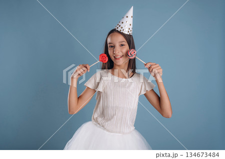 Birthday hat, with sweet candies. Young girl in white dress is against background Birthday hat, with sweet candies. Young girl in white dress is against background 134673484