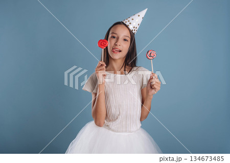 Birthday hat, with sweet candies. Young girl in white dress is against background Birthday hat, with sweet candies. Young girl in white dress is against background 134673485