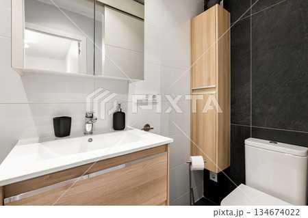 Modern bathroom with a sleek design. Features a white sink, wooden cabinetry, a mirror, and a toilet. The walls are tiled in black and white. 134674022