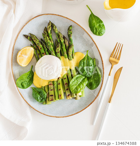 Poached egg with grilled asparagus, spinach, and hollandaise sauce on white table. Top view 134674388