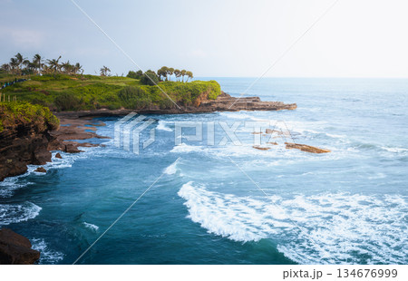 Tanah Lot coastline dramatic ocean cliffs and tropical shore near Bali temple area create calm travel mood with blue water, volcanic rocks, Bali coastal nature and open horizon under soft daylight 134676999