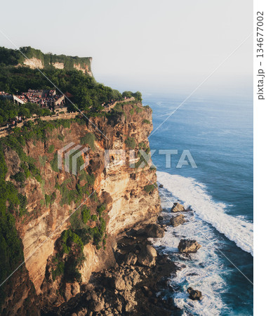 Uluwatu cliff temple Bali rising above Indian Ocean, dramatic limestone coastline with tourists at viewpoint, sacred landmark, tropical travel destination, ocean waves, spiritual atmosphere at sunset 134677002